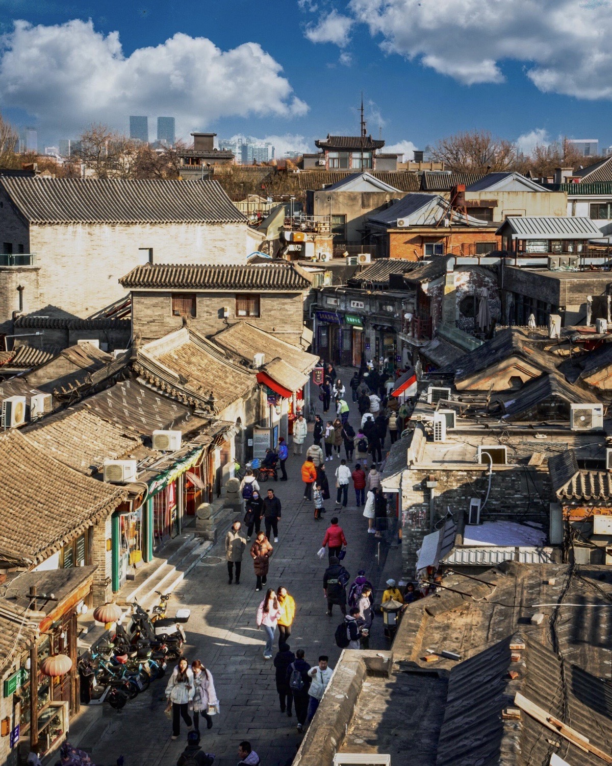 People move through a Beijing hutong beneath tiled rooftops and the modern skyline